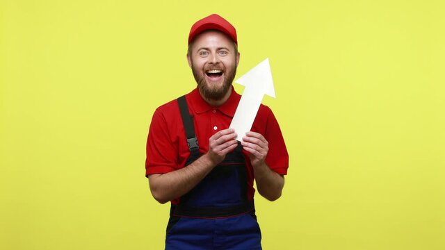 Extremely happy worker showing white paper arrow pointing up, showing increase of orders and salary, wearing blue uniform, red T-shirt and visor cap. Indoor studio shot isolated over yellow background