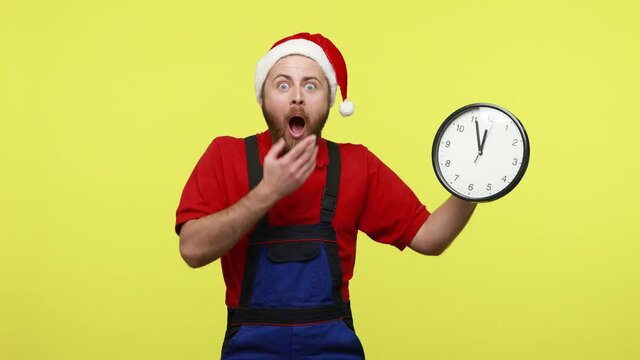 Shocked worker showing wall clock and having nervous expression, not fished work till Christmas, wearing blue overalls and santa claus hat. Indoor studio shot isolated over yellow background.