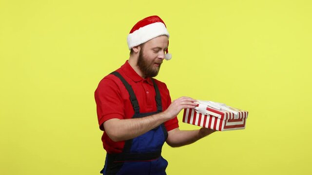 Side view portrait of handyman standing wrapped gift box in hands, looking inside and feels sadness, wearing blue overalls and santa claus hat. Indoor studio shot isolated over yellow background.