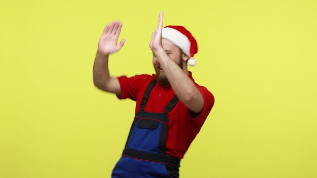 Extremely happy optimistic worker dancing on corporate Christmas party, being in excellent mood, wearing blue overalls and santa claus hat. Indoor studio shot isolated over yellow background.