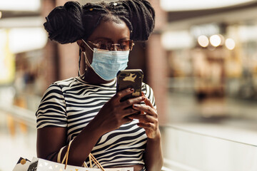 african woman, wearing a medical protective mask on her face, with shopping bags, using phone