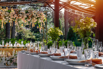 Wedding table setting decorated with fresh flowers in a brass vase. Wedding floristry. Banquet table for guests outdoors with a view of green nature at sunset.. Bouquet with roses