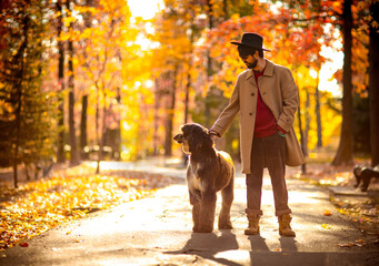 a handsome, brutal man in a coat and hat, walking in the park during the day, in autumn, with a large thoroughbred dog