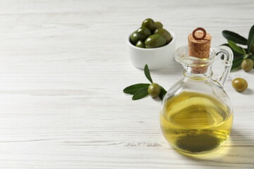 Glass jug of oil, ripe olives and green leaves on white wooden table. Space for text