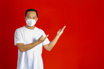 Asian man wearing a medical mask showing his hand to copy the space on his side on a red background