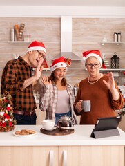 Happy family wearing santa hat greeting remote parents enjoying christmas holiday together during online videocall meeting conference in xmas decorated kitchen. Child celebrating winter season