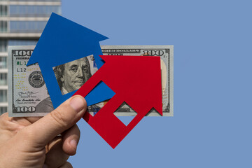 Symbolic paper houses and a 100 american dollar bill against the backdrop of a modern high-rise building and blue sky