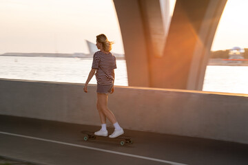 Woman skateboarder riding longboard on street at sunset. Female longboarder practicing alone. Casual active girl skater training or relaxing after work on skateboard skating alongside river in city