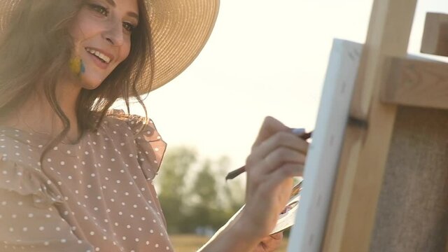 Close-up of young Artist stained with paints in big hat stands on field in rays of setting sun and draws with brushes on canvas on an easel. he holds brushes in his teeth. Painter draws in nature.