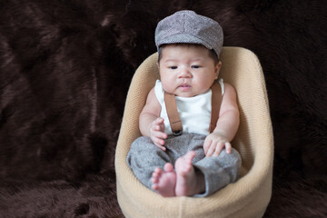A newborn boy wearing a casual white shirt and hat with gray pants is sitting on a baby sofa on a brown background.