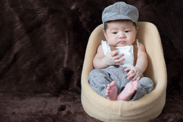 A newborn boy wearing a casual white shirt and hat with gray pants is sitting on a baby sofa on a brown background.