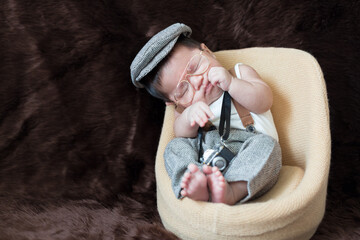 A newborn boy wearing a casual white shirt and hat with gray pants is sitting on a baby sofa on a brown background.