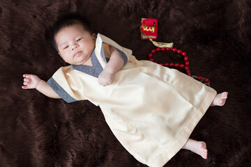 A cute Asian infant Muslim in thobe and rosary beads on red, muslim bead on bed in the home, translation is the name of muslim God "Allah" in arabic text