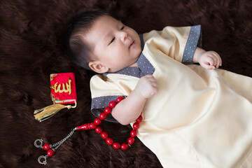 A cute Asian infant Muslim in thobe and rosary beads on red, muslim bead on bed in the home, translation is the name of muslim God "Allah" in arabic text
