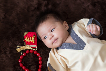 A cute Asian infant Muslim in thobe and rosary beads on red, muslim bead on bed in the home, translation is the name of muslim God "Allah" in arabic text