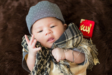 A cute Asian infant Muslim in thobe with prayer cap on bed in home, translation is noble quran