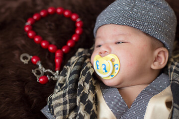 A cute Asian infant Muslim in thobe with prayer cap on bed in home, translation is noble quran