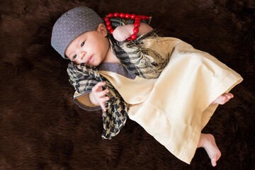 A cute Asian infant Muslim in thobe with prayer cap on bed in home.