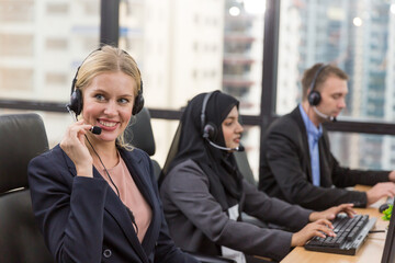 Smiling female customer support operator with headset in office. Team of diversity call center or...