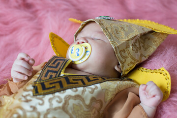 A newborn baby wearing a Chinese emperor's costume gold colour lying on a pink blanket.