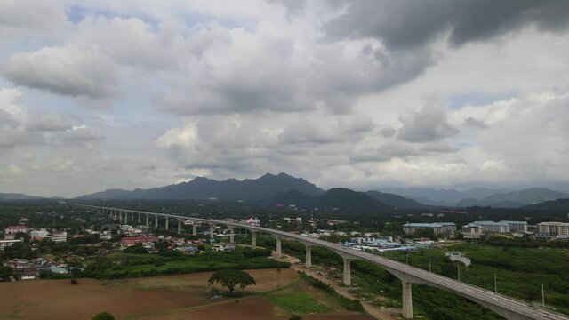 Aerial footage towards as town, Muak Klek, in Saraburi, Thailand, revealing an elevated railway on the right and mountains in the horizon with rain clouds.
