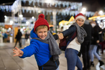 Portrait of tween boy pointing to desired thing and pulling mothers hand during walk at festive fair on Christmas Eve