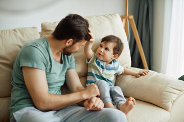 Small boy touches father's hair while relaxing with him on sofa at home.