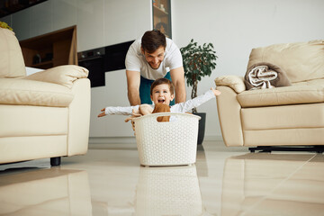Happy kid with arms outstretched has fun while father pushes him in laundry basket at home.