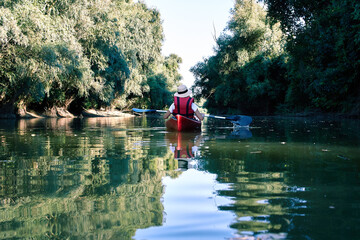 Back view on woman paddles red kayak on river or lake near green trees at summer