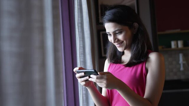 A young Indian cheerful attractive woman is holding a mobile phone in hands and watching a comedy video or movie on an online stream or OTT platform or smiling.Asian smiling woman using smartphone.
