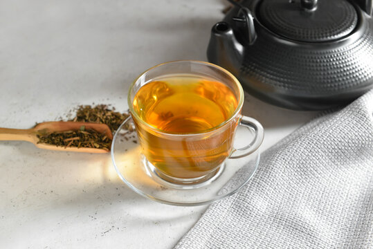 Cup of tasty hojicha green tea, pot and spoon on light background