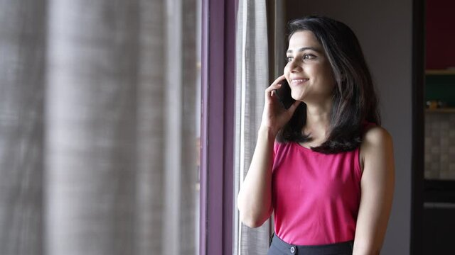 Indian young woman talking on smartphone with her family and friends.Closeup shot of a beautiful Asian girl happily talking to her friend on a phone call during standing near a window.
