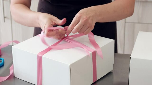 Woman pastry chef packs cake in white box and ties up with pink ribbon, close-up. Slow motion. Preparing for delivery