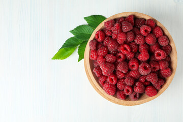 Red juicy raspberries in a wooden bowl on a white wooden background