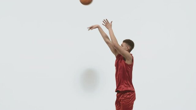 The basketball guy throws the ball into the basket and is glad that he scored. Young sportsman in sports uniform trains on a white studio background. Slow motion.