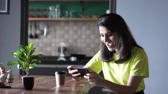 Portrait of a indian woman hand holing mobile phone sitting on a table playing a video game together with an excited and happy expression. Smiling woman having fun and enjoying in free time.