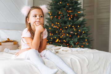 Obraz na płótnie Canvas Cute sad funny little girl in pink dress with present gift box sitting on bed in room with Christmas tree. Portrait of unhappy child in cottage house in christmas time, happy New Year