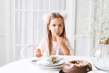 Obraz na płótnie Canvas Funny cute little girl with long hair in light pink dress holding popcake in hands on festive table in bright living room at home. Christmas time, birthday girl