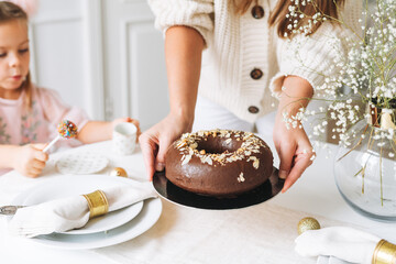 Young woman mother in white holding in hands Large round chocolate almond cake on the table with...