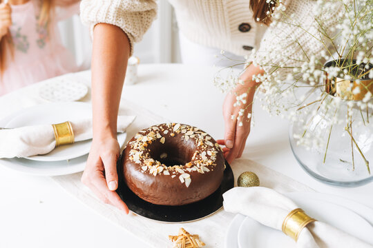 Young woman mother in white holding in hands Large round chocolate almond cake on the table with New Year serving, christmas white scandinavian festive table, family event