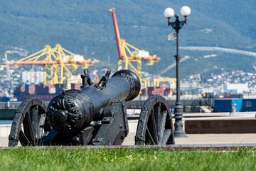 Embankment of Admiral Serebryakov. Old ship's cannon is pointed towards sea. Coast of Tsemesskaya Bay. Blurred background of harbor cranes at seaport. Novorossiysk, Russia - September 15, 2021