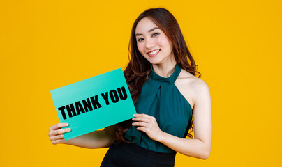 Young happy Asian women smiling and holding green cardboard sign with the word Thank you in front of her. Standing isolated on bright yellow background