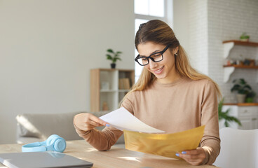 Smiling young woman who is excited by good news reads letter unfolded from paper envelope. Excited...