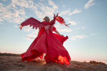 A plump blonde middle-aged woman with red angle wings in a desert with dunes and sand in a nice summer or spring sunny day with blue sky
