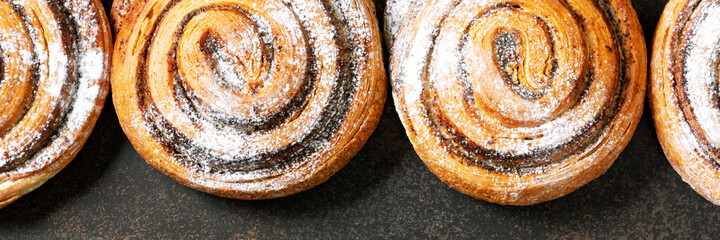 A sweet spiral bun with poppy seeds and powdered sugar of a large size on a dark background. Delicious yeast pastries top view. The concept of home-made high-calorie baking. Banner