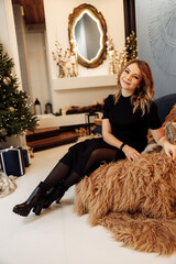 Stylish gorgeous woman sitting in cozy chair near decorated Christmas tree. Beautiful female in fashion clothes posing, looking directly at the camera, spending winter holidays at home, xmas concept