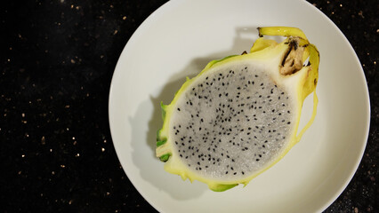 Close up of half a yellow dragon fruit, with the flesh visible, served on a white plate. Top view, with copy space on the left.