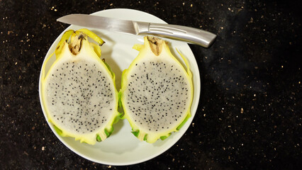Flat lay of a yellow dragon fruit cut into half, served on a white plate, with a knife placed at the side.