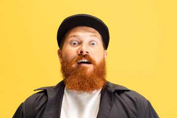 Close-up portrait of stylish red-bearded man, barber in black cap looking at camera isolated over yellow background. Concept of emotions, feelings.