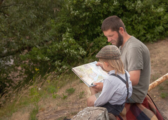 Fototapeta premium a father and son in vintage clothes are sitting by the road and looking at a map and discussing a tourist route